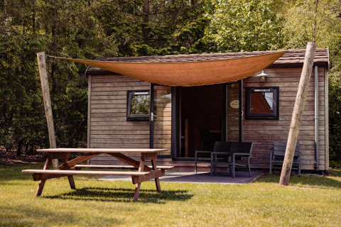 Photo de la Woodlodge au Camping the Noetselerberg, Pays-Bas, avec un lodge en bois, table de pique-nique et voile d'ombre.