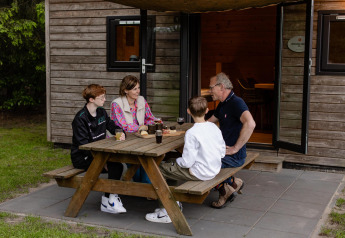 Een gezin zit aan een picknicktafel voor een Woodlodge op Camping de Noetselerberg in Nederland.