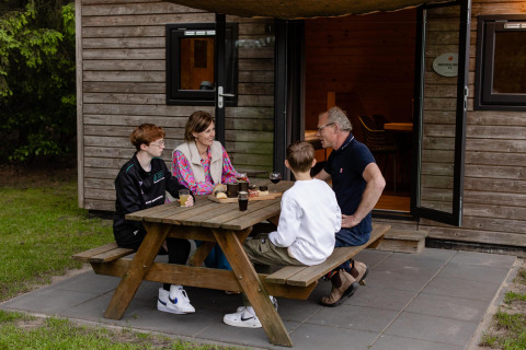 Een gezin zit aan een picknicktafel voor een Woodlodge op Camping de Noetselerberg in Nederland.