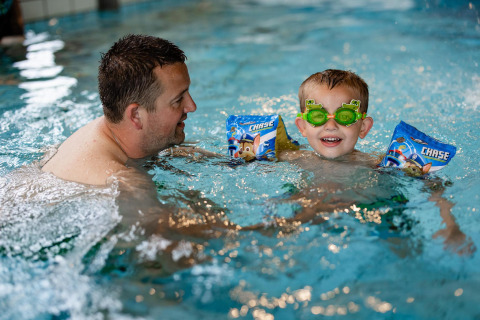 Erwachsener und Kind spielen lachend im Schwimmbad eines Glamping-Resorts, das Kind trägt Schwimmflügel.