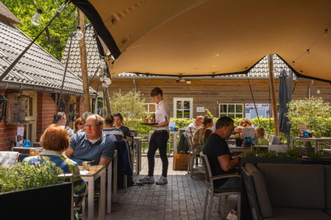 Terrasse de restaurant sur un site de glamping, clients mangeant sous un auvent lors d'une journée ensoleillée.