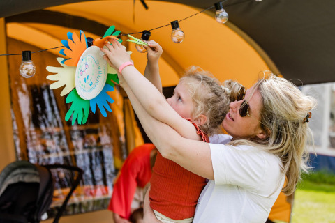 Une femme aide une enfant à accrocher des décorations colorées sous une tente de glamping bien éclairée.
