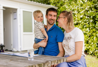 Een familie zit buiten aan een houten tafel bij een glampingverblijf, omringd door veel groen.