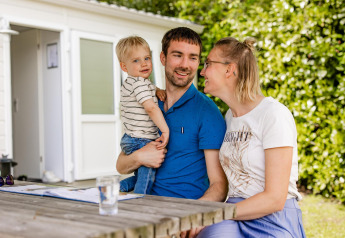 Een gezin zit aan een houten tafel buiten bij een glamping-accommodatie, omgeven door groen.