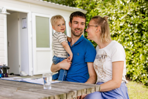 Een familie zit buiten aan een houten tafel bij een glampingverblijf, omringd door veel groen.