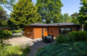Hyggelig lodge i træ med terrasse, havebord og masser af grønne træer, Heide lodge i Bungalowpark Het Verscholen Dorp.