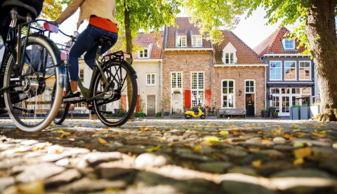 Dos personas en bicicleta pasan por casas pintorescas y árboles verdes en Heide lodge de Bungalowpark Het Verscholen Dorp.