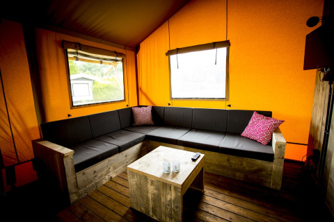 Vista interior de una tienda safari Glamping Lodge con sofá esquinero, mesa de madera y paredes naranjas.