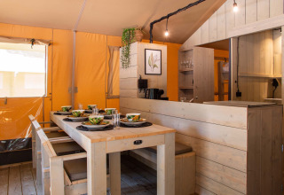 Mesa de comedor y cocina dentro de una tienda safari en Holiday Park Gelloo, Países Bajos, con interior de madera.