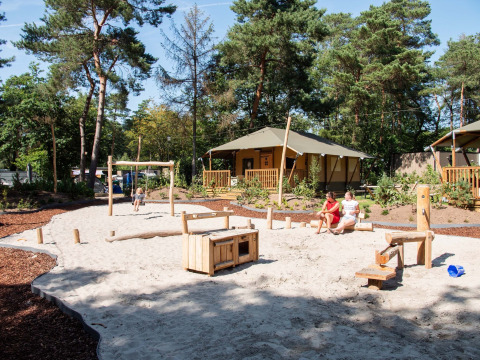 Tente safari au Holiday Park Gelloo, Pays-Bas, avec aire de jeux sablée et familles, entouré de forêt.