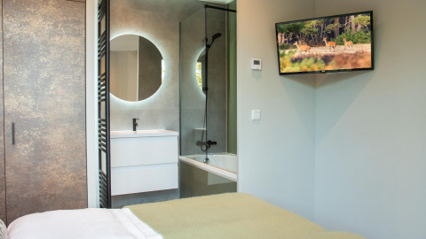 Habitación moderna de lodge con cama cómoda, televisor en la pared y baño con espejo redondo y ducha.