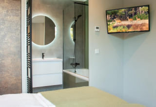 Habitación moderna de lodge con cama cómoda, televisor en la pared y baño con espejo redondo y ducha.