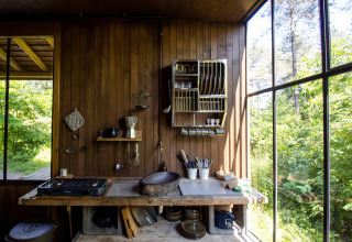 Cocina rústica de cabaña con paredes de madera, grandes ventanas y vista a la exuberante naturaleza exterior.