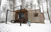 Cabaña moderna de madera en un bosque nevado con terraza, asador y luces exteriores en invierno.