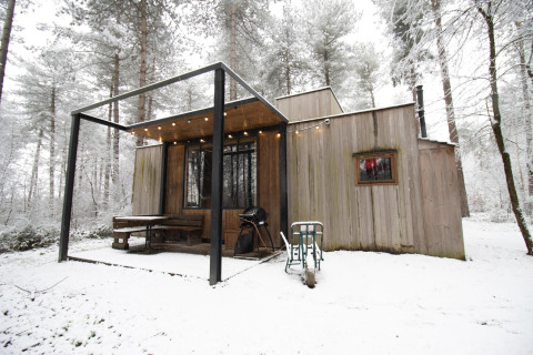 A modern wooden cabin in a snowy forest setting with a terrace, grill, and string lights in winter.