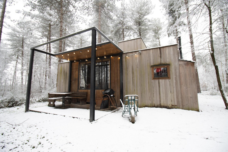 Moderne houten hut in een besneeuwd bos met terras, barbecue en sfeerverlichting in de winter.