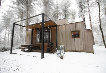 Eine moderne Holzhütte im verschneiten Wald mit Terrasse, Grill und Lichterkette im Winter.