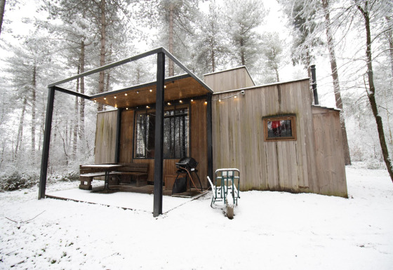 A modern wooden cabin in a snowy forest setting with a terrace, grill, and string lights in winter.