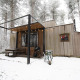 Eine moderne Holzhütte im verschneiten Wald mit Terrasse, Grill und Lichterkette im Winter.