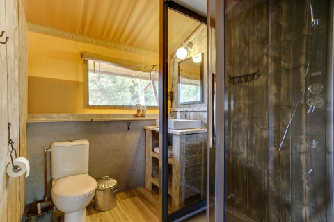 Baño en tienda safari con inodoro, lavabo, ducha de madera rústica y ventana grande con vista natural.