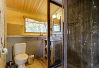 Badkamer in safaritent met toilet, wastafel, houten douche, groot raam en natuurlijke houtaccenten.