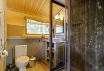 Baño en tienda safari con inodoro, lavabo, ducha de madera rústica y ventana grande con vista natural.