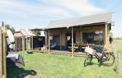Alojamiento de glamping moderno con porche cubierto, bicicleta y zona de descanso sobre césped soleado.