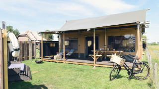 A modern glamping lodge with a covered porch, bicycle, and outdoor seating area on a sunny grassy field.