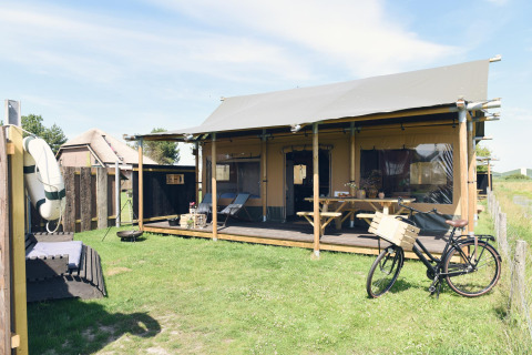 Luxe glampingaccommodatie met overdekte veranda, fiets en zitruimte op een zonnig grasveld.