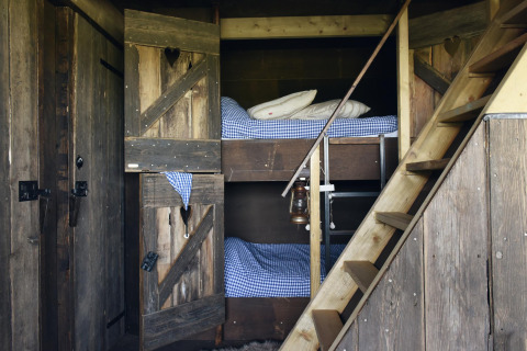 Accogliente stanza glamping con letti a castello in legno, biancheria a quadretti e scala rustica.