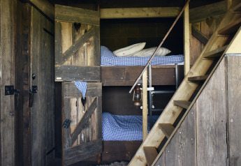 Accogliente stanza glamping con letti a castello in legno, biancheria a quadretti e scala rustica.