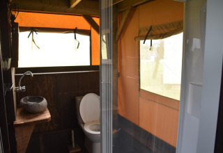 Baño en una cabaña con inodoro, lavabo de piedra y ducha, rodeado de paredes tipo tienda de campaña.
