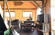 Interior view of a glamping tent with wooden furniture, a sofa, dining table, crates, and natural light.