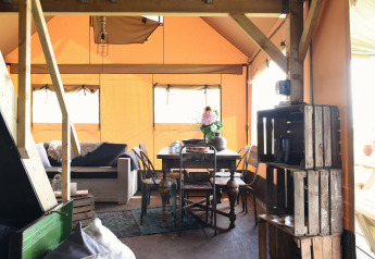 Vista interior de una tienda glamping con muebles de madera, sofá, mesa de comedor y luz natural.