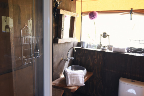 Baño rústico en glamping con paredes de madera, lavabo de piedra, toallas y ventana con mucha luz.