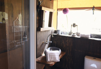 Baño rústico en glamping con paredes de madera, lavabo de piedra, toallas y ventana con mucha luz.