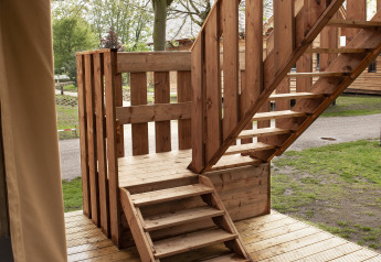 Escalera y balcón de madera junto a una tienda safari en Forest Lodge Junior, Sandberghe, Países Bajos, rodeado de árboles.