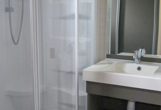 Un baño moderno en un albergue con ducha de cristal, lavabo blanco con grifo y un gran espejo iluminado.