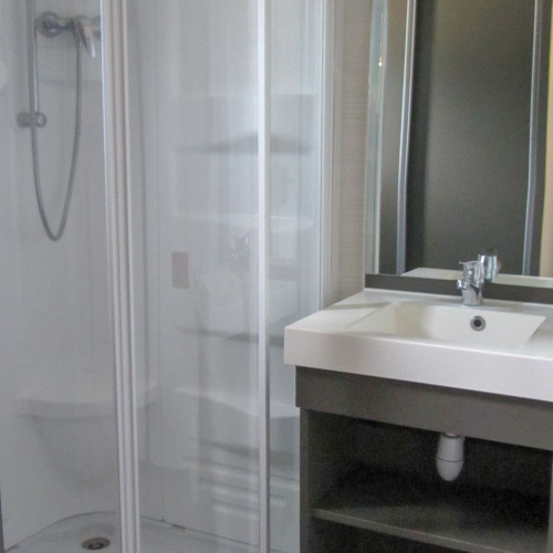 Un baño moderno en un albergue con ducha de cristal, lavabo blanco con grifo y un gran espejo iluminado.