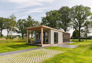 Modern tiny house in a green field with a large covered patio, surrounded by grass and tall trees.