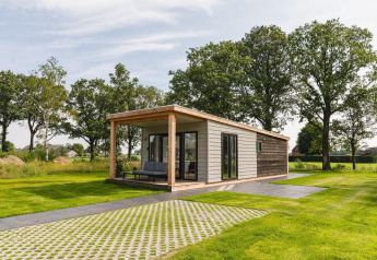 Moderne tiny house met groot overdekt terras op een groene weide, omgeven door hoge bomen.