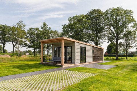 Modern tiny house in a green field with a large covered patio, surrounded by grass and tall trees.
