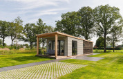 Moderne tiny house op een groen veld met overdekt terras, omgeven door gras en hoge bomen.