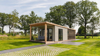 Modern tiny house in a green field with a large covered patio, surrounded by grass and tall trees.