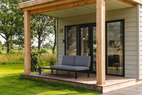 Tiny house con veranda coperta, moderno divano grigio e vista verde su un tranquillo paesaggio rurale.