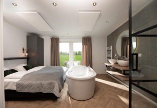 Modern tiny house bedroom featuring a freestanding bathtub, double bed, and view of green fields outside.