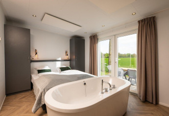 Bright tiny house bedroom featuring a double bed, freestanding bathtub, and views of nature through glass doors.
