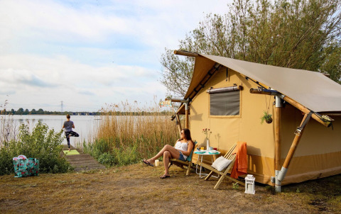 Mujer relajándose frente a una tienda safari junto al lago, mientras otra hace yoga en Marina Parcs Almere, Países Bajos.