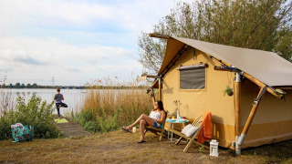 Mujer relajándose frente a una tienda safari junto al lago, mientras otra hace yoga en Marina Parcs Almere, Países Bajos.