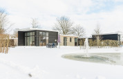 Moderne lodge med store vinduer, snedækket landskab og springvand ved Vakantiepark Hölte, Nederlandene.